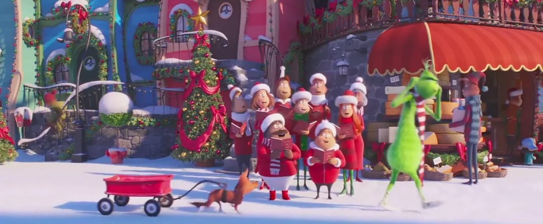A group of festive Whos sing carols outside colorful, snow-covered houses decorated for Christmas. The Grinch, dressed in green, walks by with a brown dog and a red wagon in the snowy scene.