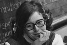 A young woman with glasses and pigtails sits in front of a chalkboard, resting her chin on her hand and smiling slightly. She wears a sleeveless sweater over a long-sleeved shirt. Chalk writing is visible on the board behind her.