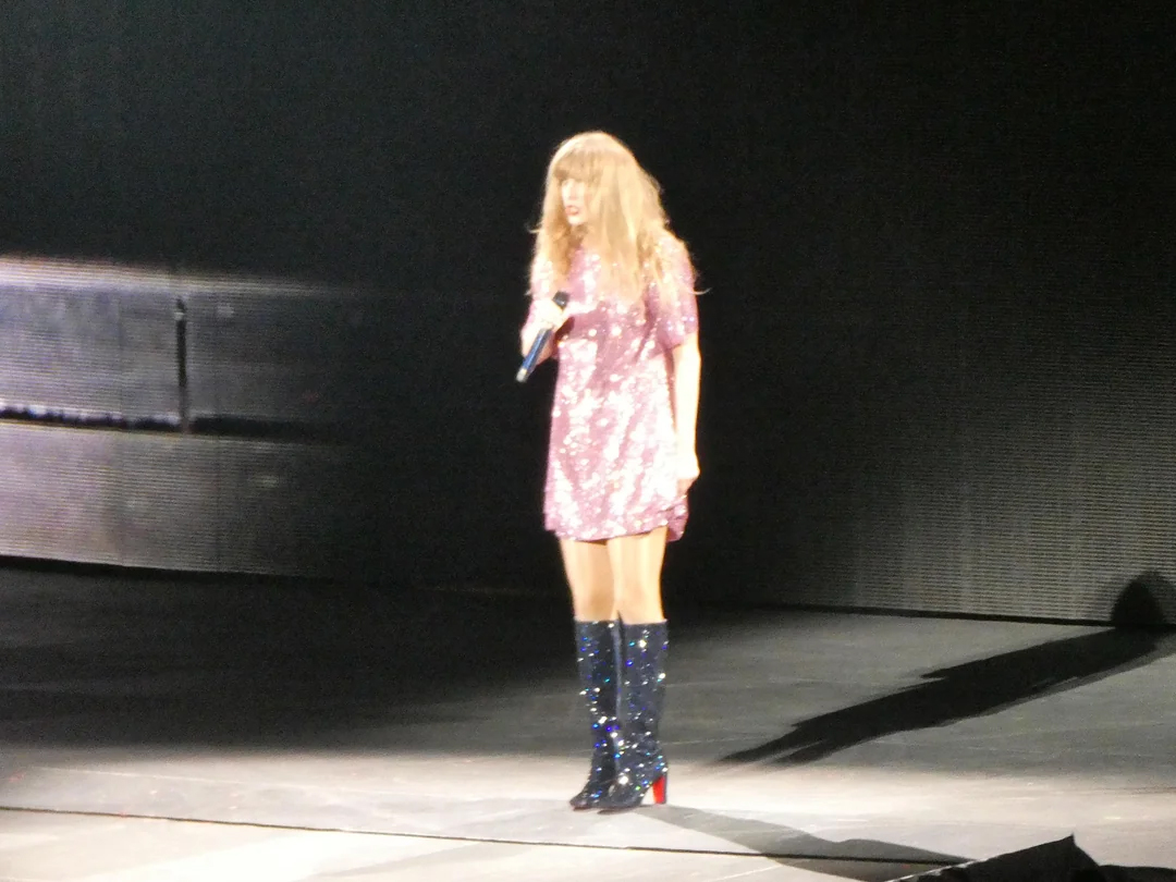 A woman with long blonde hair in a sparkly pink dress and tall black boots stands onstage holding a microphone, with a dark background behind her.
