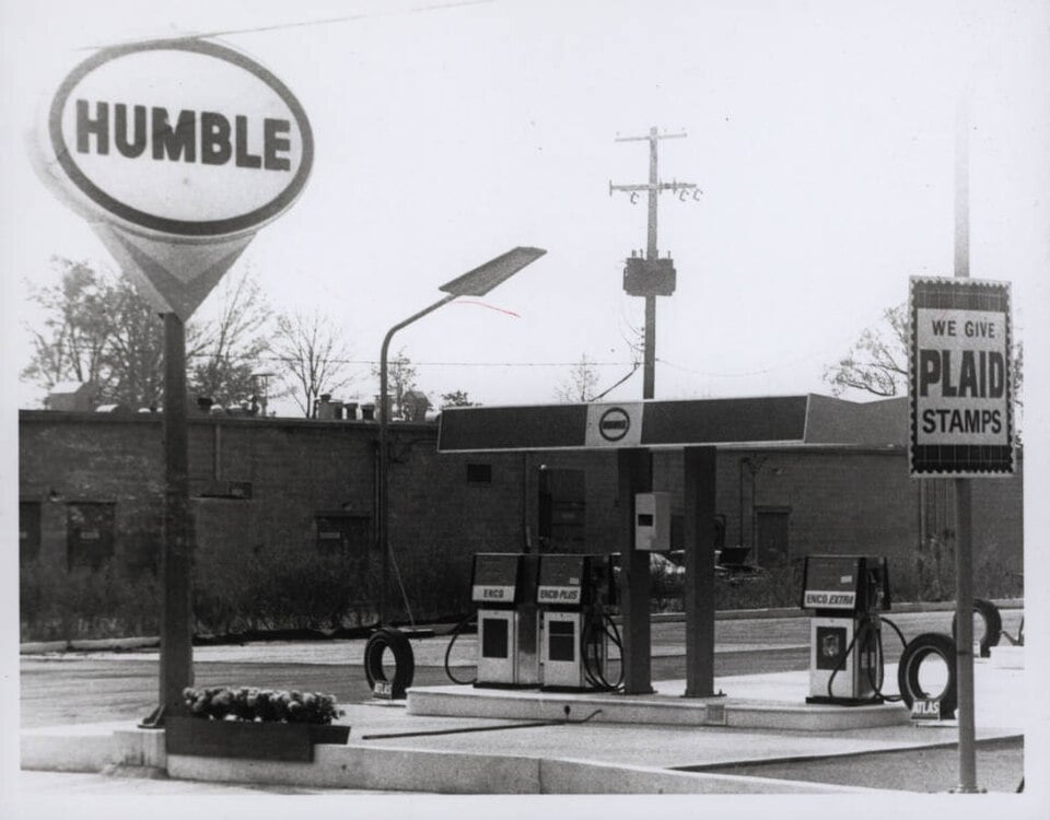 Black and white photo of a vintage Humble gas station with two gas pumps, a large Humble sign, and a sign that reads "We give Plaid Stamps." A brick building and power lines are in the background.