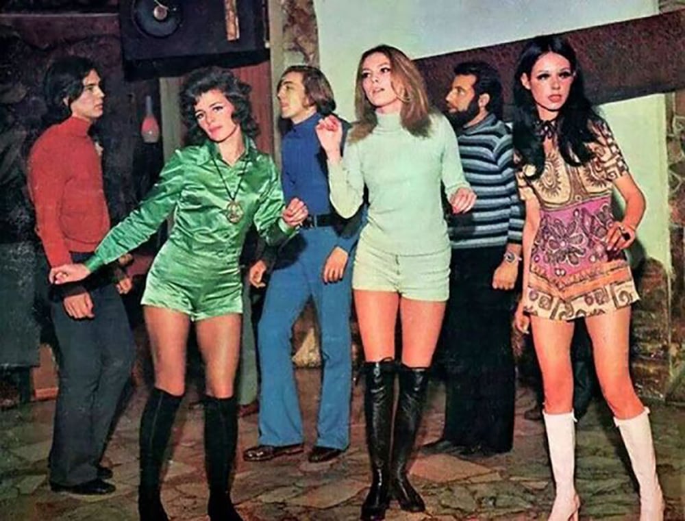 A group of young adults in colorful 1970s fashion dance and socialize indoors, with women in mini dresses and boots, and men in flared pants and shirts, creating a lively retro atmosphere.