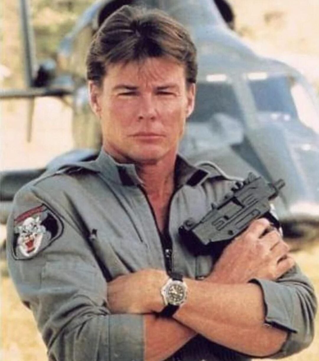 A man in a gray flight suit stands with arms crossed, holding a black firearm. He has short brown hair and a serious expression. A helicopter is blurred in the background. There is a patch with a wolf on his sleeve and a watch on his wrist.