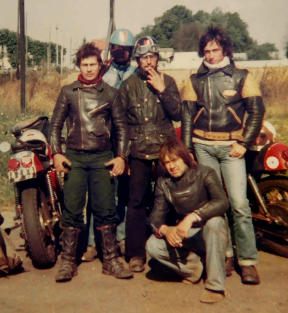 Five bikers wearing black leather jackets pose together outdoors beside two motorcycles, with trees and a grassy area in the background. Four are standing, and one is crouching in front.
