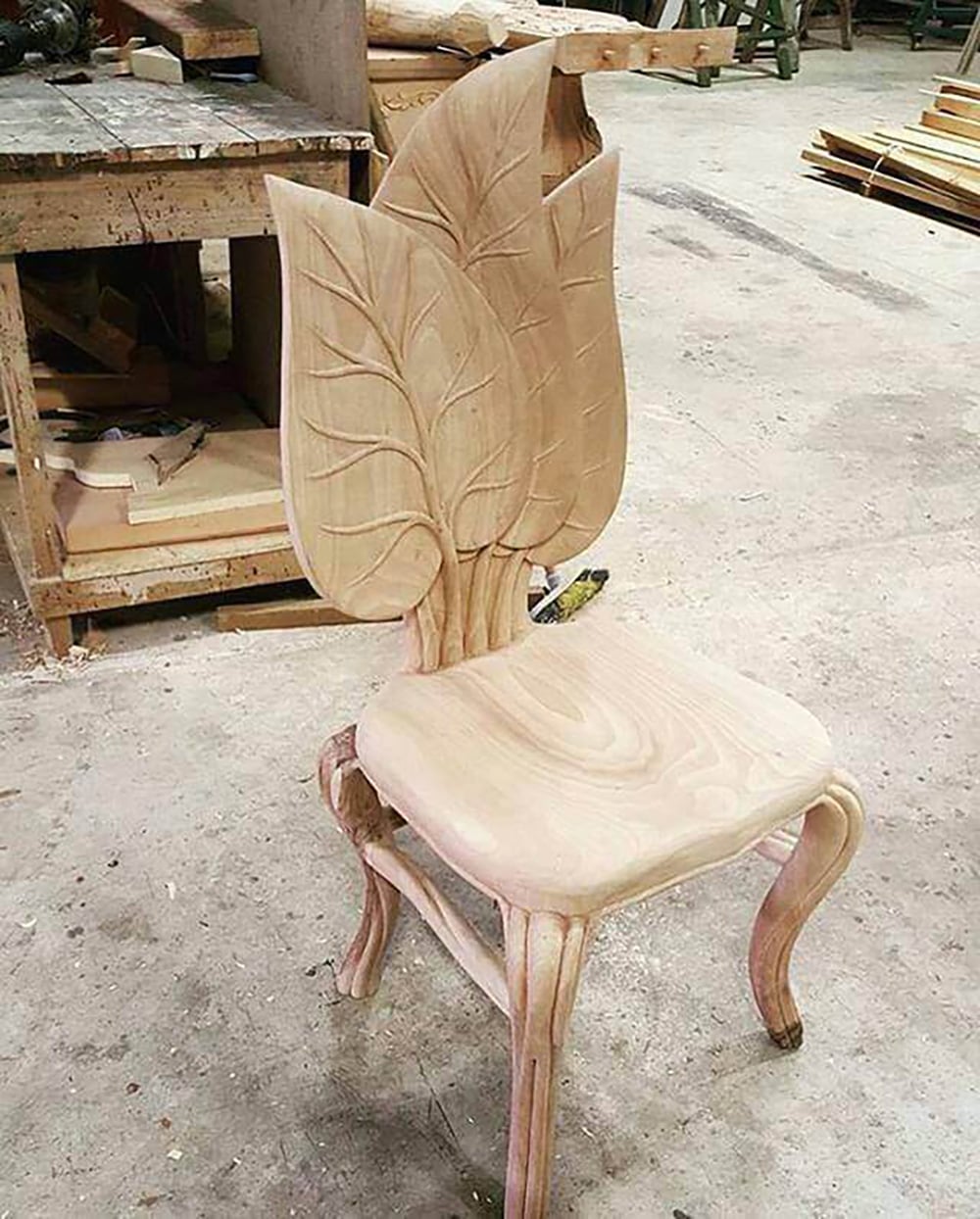 A wooden chair with a backrest carved to resemble three large, overlapping leaves, and curved legs, sits in a workshop with wood and tools in the background.