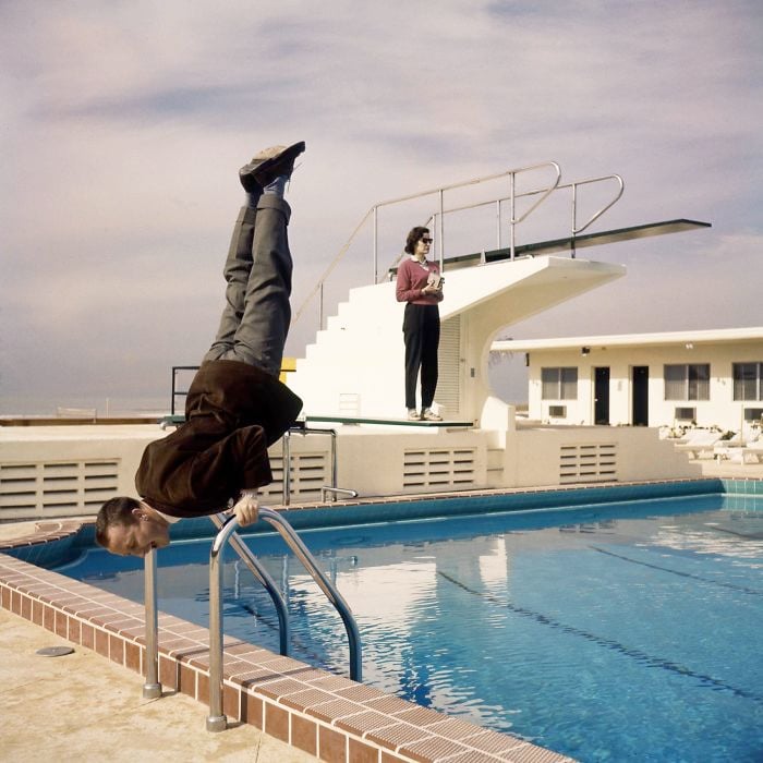 A man balances upside down on a pool ladder by a swimming pool, while another person stands on a diving board in the background, holding a towel. The sky is partly cloudy.