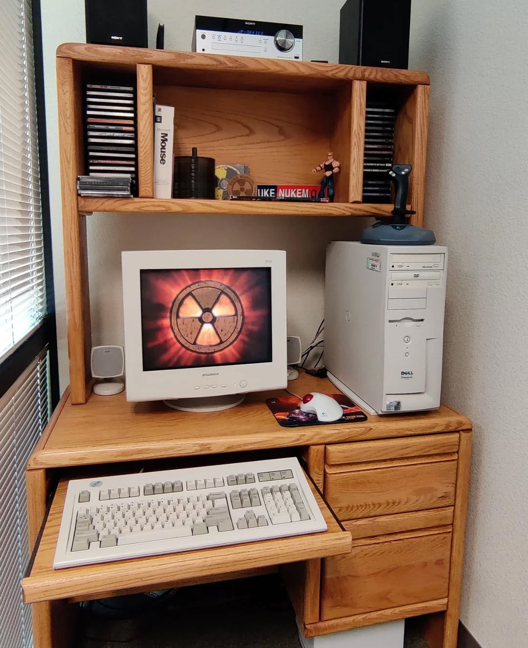 A vintage desktop computer setup on a wooden desk, featuring a CRT monitor displaying a radioactive symbol, a large Dell tower, speakers, a keyboard, mouse, CDs, books, and a figurine on the shelf above.