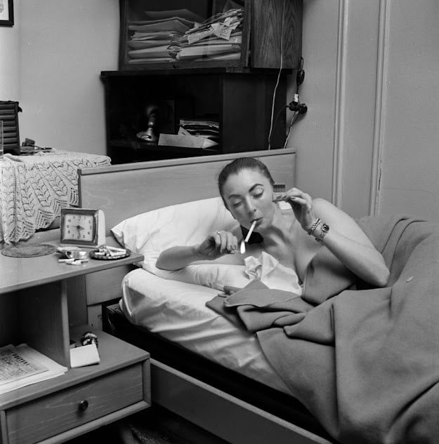 A woman sits up in bed, lighting a cigarette with a match. Papers and a telephone are stacked on a shelf behind her, and there is a nightstand with a clock and other items beside the bed.