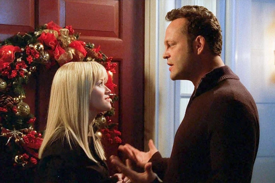 A woman and man stand facing each other in front of a red door decorated with a Christmas wreath, engaged in conversation in a warmly lit room.