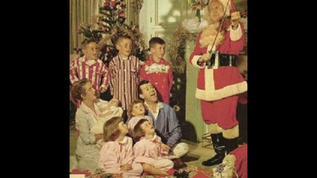 A group of children and adults in pajamas sit around a Christmas tree, watching someone dressed as Santa Claus play the violin in a festive living room.