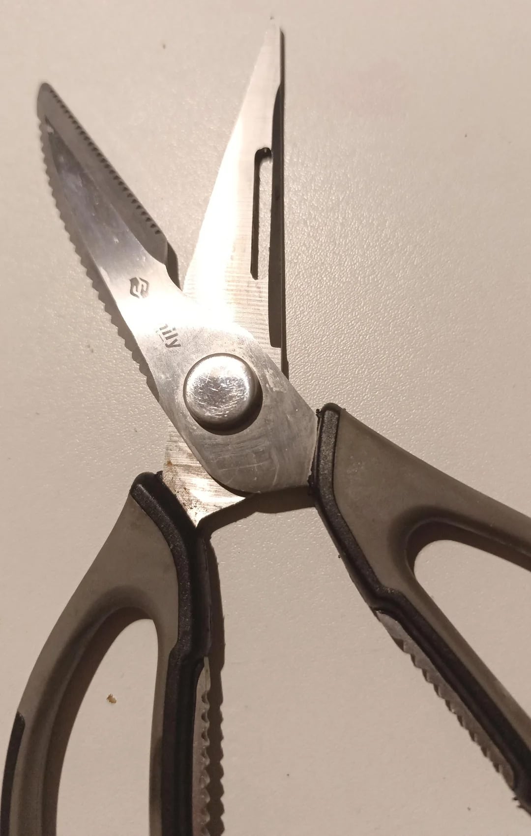 A close-up of a pair of kitchen scissors with black and gray handles, open and resting on a white surface. The blades have a serrated edge and a notch for cutting tougher materials.