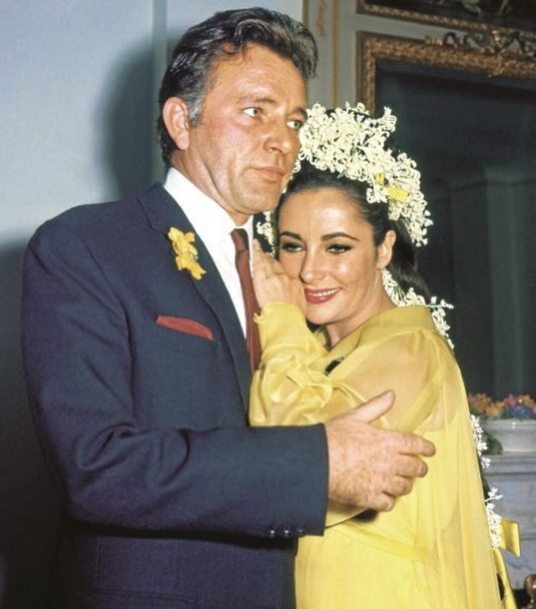A man in a dark suit embraces a smiling woman in a yellow dress with white floral headpieces. The woman leans affectionately against his chest; both appear happy and dressed for a special occasion.