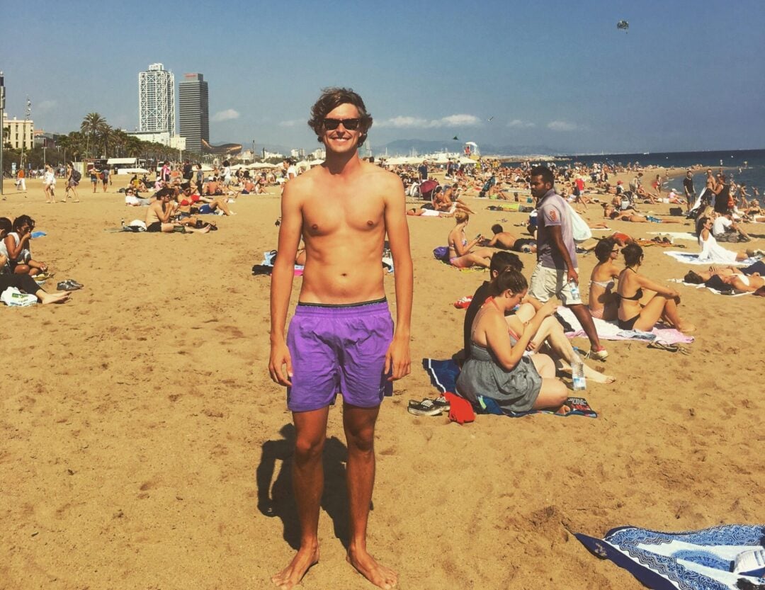 A young man wearing purple swim trunks stands and smiles on a crowded sandy beach under a clear blue sky, with many people sunbathing and relaxing around him; city buildings are visible in the background.