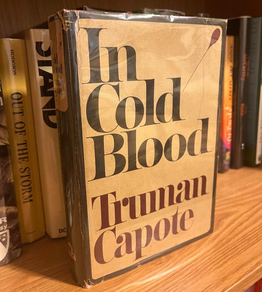 A hardcover copy of "In Cold Blood" by Truman Capote is displayed on a wooden bookshelf, surrounded by other books. The book’s dust jacket is slightly worn and protected by a clear cover.