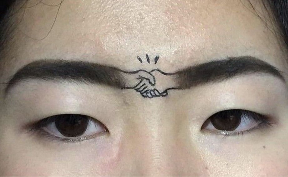 A close-up of a person’s face showing their eyebrows, with a drawing of two hands shaking tattooed or drawn above the nose, making the eyebrows appear connected by the handshake.