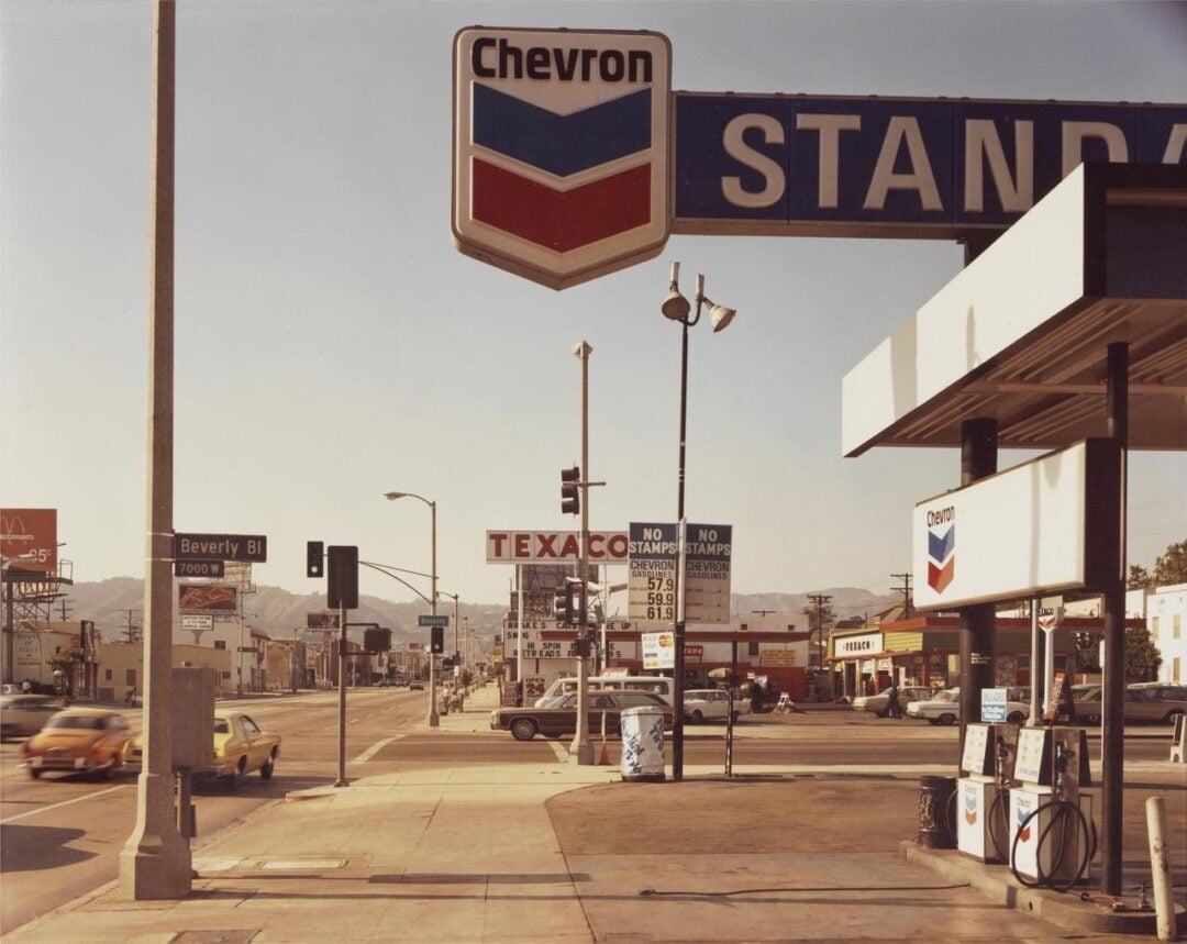 A vintage photo of a Chevron gas station on a sunny street corner, with signs for Texaco and Standard. Cars are driving by, and a McDonald's sign is visible in the background. The scene has a retro, mid-century feel.