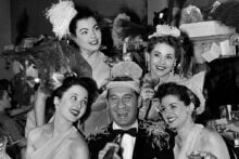 A man in a suit and party hat sits at a table surrounded by four women in festive dresses and hats, all smiling and holding drinks, in a lively, decorated backstage dressing room.