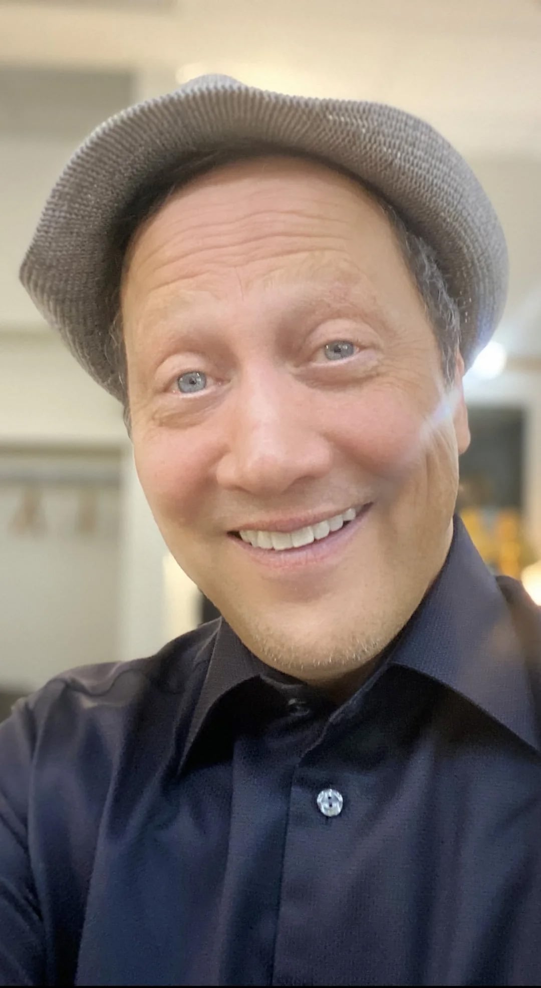 A smiling man wearing a gray hat and a dark shirt takes a selfie indoors, with a blurred background and soft lighting.