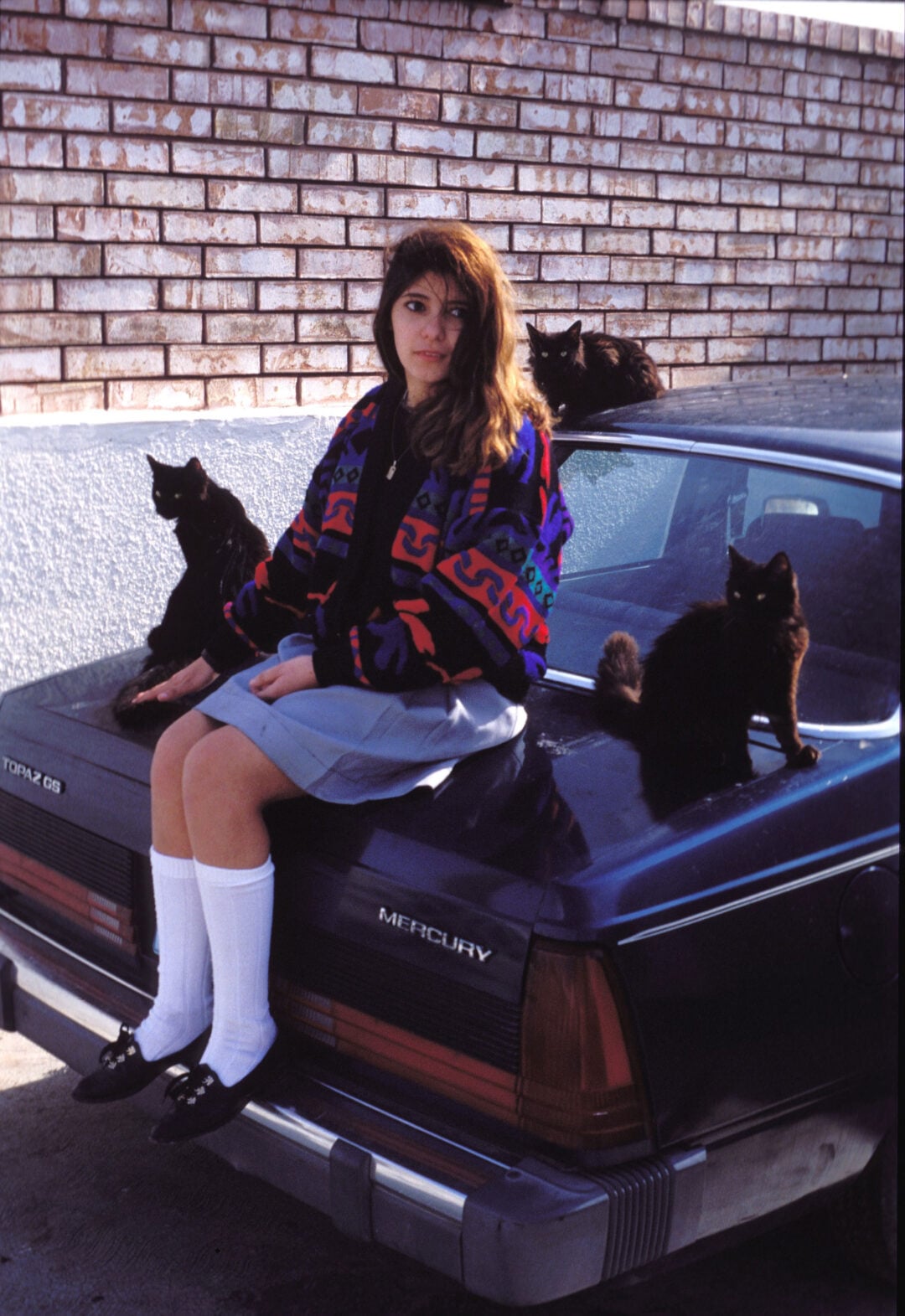 A young woman sits on the trunk of a dark blue Mercury car, surrounded by four black cats. She wears a colorful sweater, blue skirt, white socks, and black shoes. A brick wall is visible in the background.
