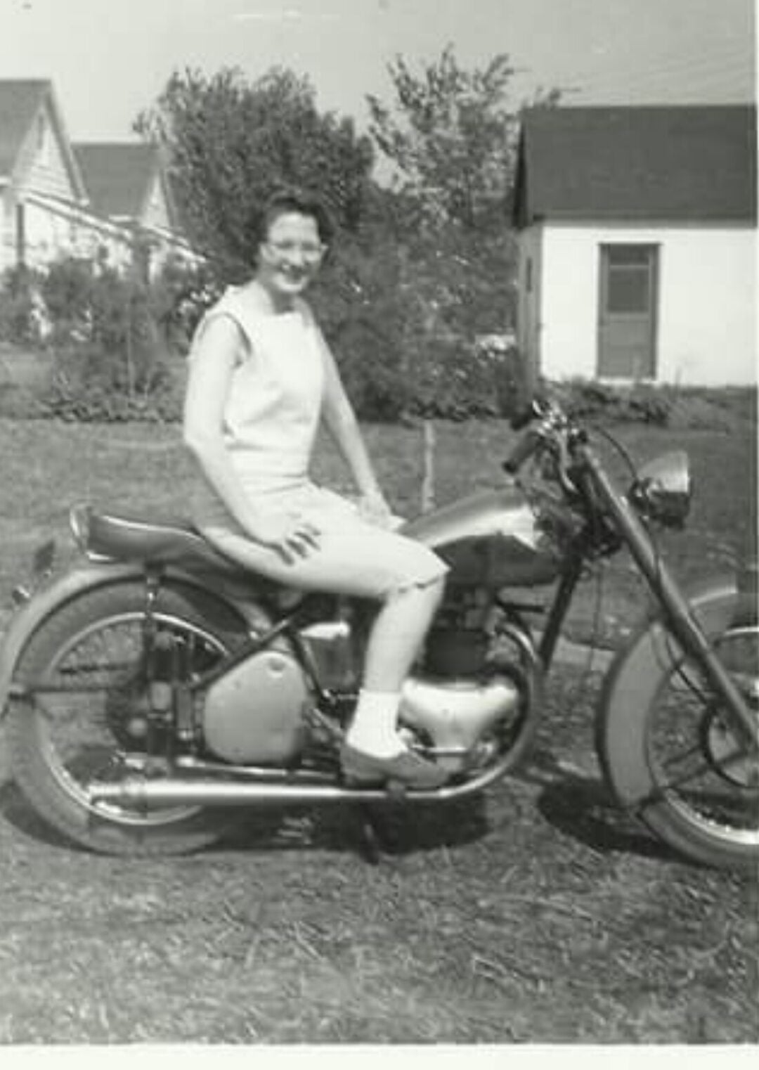 A woman in a sleeveless dress and glasses sits smiling on a vintage motorcycle in a yard, with houses and trees visible in the background. The photo is black and white.