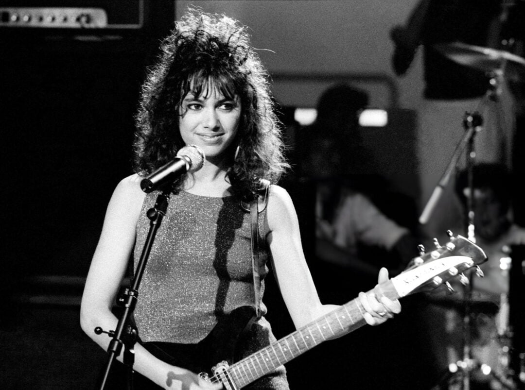 A woman with curly dark hair stands at a microphone, smiling, and holds an electric guitar on stage. She wears a sleeveless sparkly top and appears to be performing. The image is in black and white.