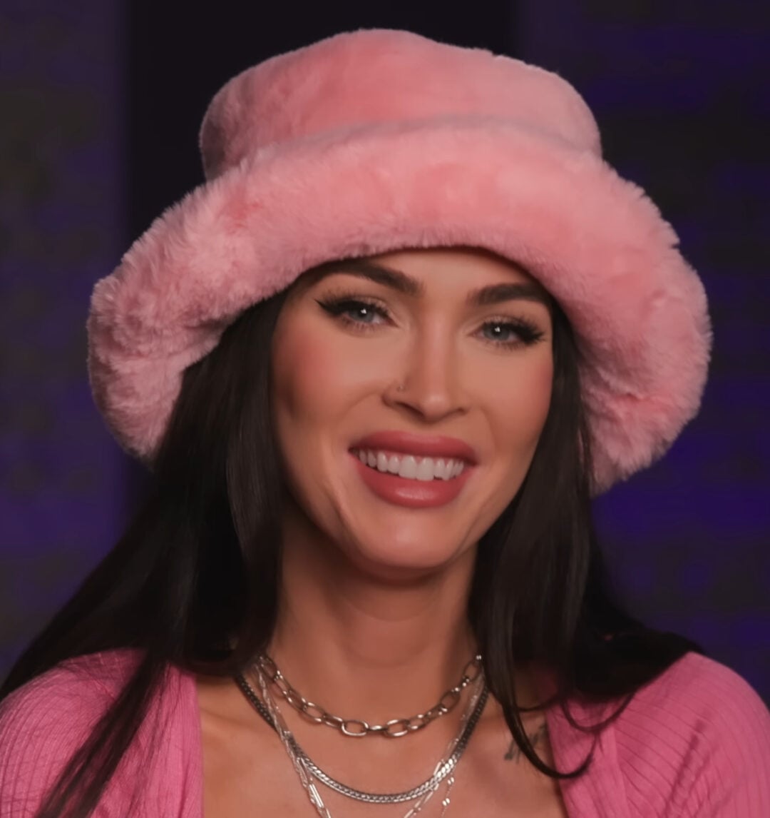 A woman with long dark hair smiles while wearing a fuzzy pink hat and a matching pink top, accessorized with layered silver necklaces, set against a softly blurred dark background.