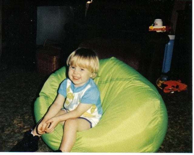 A young child with blond hair sits smiling on a bright green bean bag chair indoors, wearing a blue and white outfit with yellow designs. The background includes toys and household items.