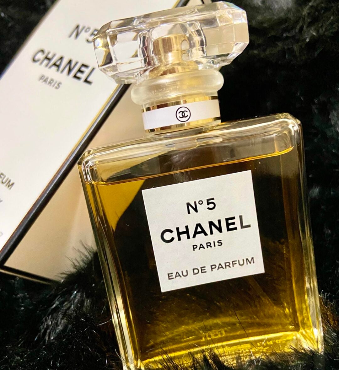 A bottle of Chanel No. 5 Eau de Parfum sits on a black textured surface, with its box positioned behind it, partially visible. The clear bottle has a gold-toned liquid and a faceted, transparent cap.