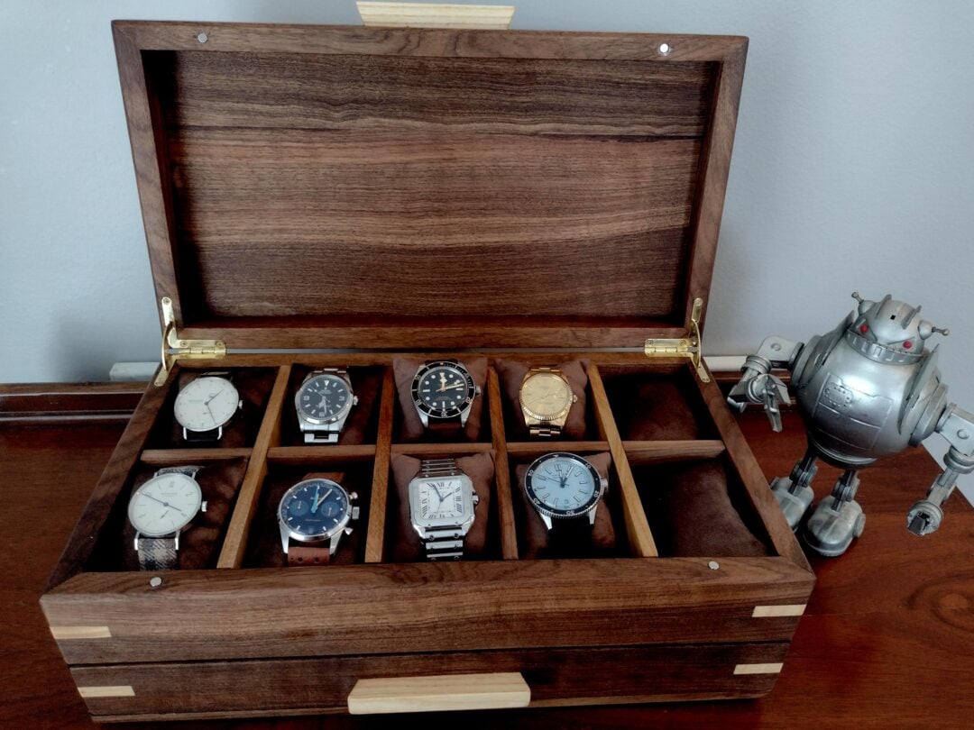 A wooden box with eight compartments holds assorted wristwatches, displayed with an open lid. A metallic toy robot stands to the right of the box on a wooden surface.
