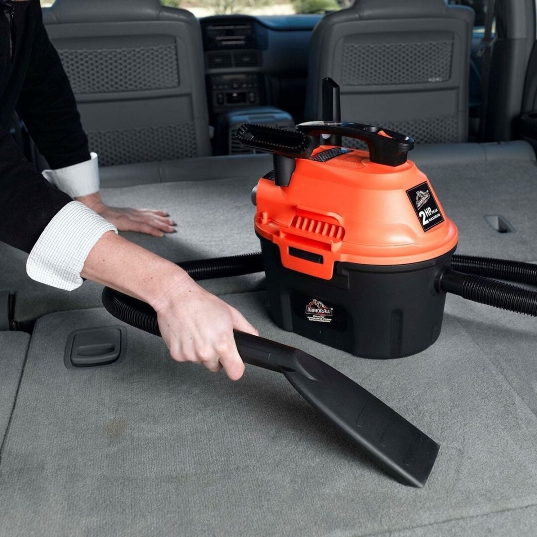 A person uses a compact, orange and black vacuum cleaner to clean the backseat area of a car with a flat nozzle attachment.