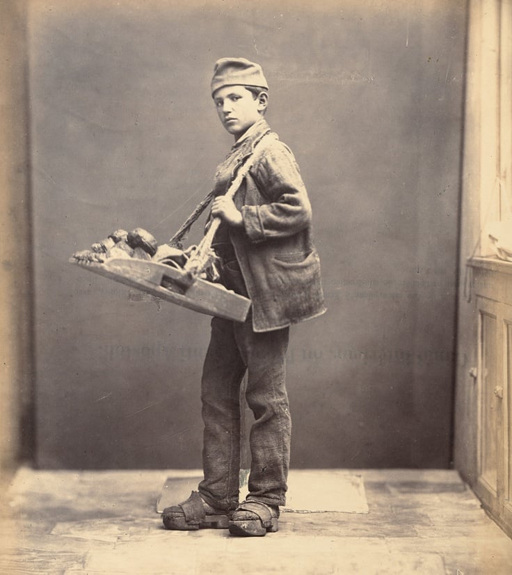 A young boy stands indoors wearing a cap, jacket, and clogs, holding a wooden tray with brushes and other tools over his shoulder, looking at the camera against a plain backdrop.