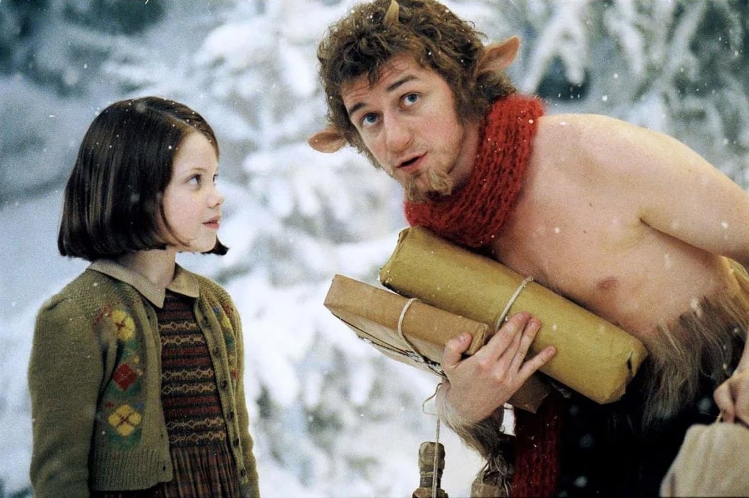 A young girl in a dress and sweater stands beside a faun with curly hair, pointy ears, and a red scarf, holding wrapped parcels in a snowy forest.
