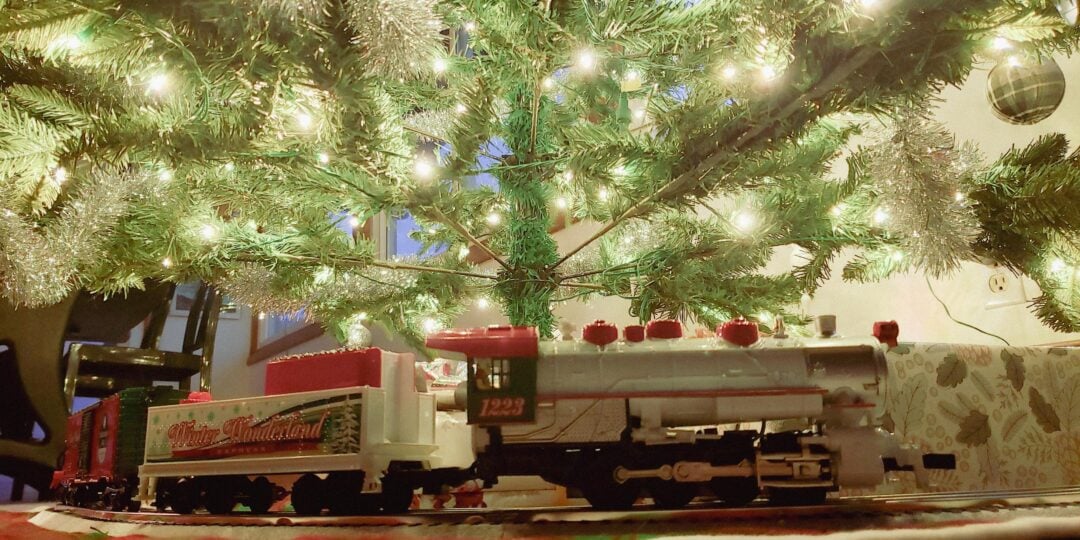 A model train set circles beneath a decorated Christmas tree glowing with white lights, creating a festive holiday scene indoors.