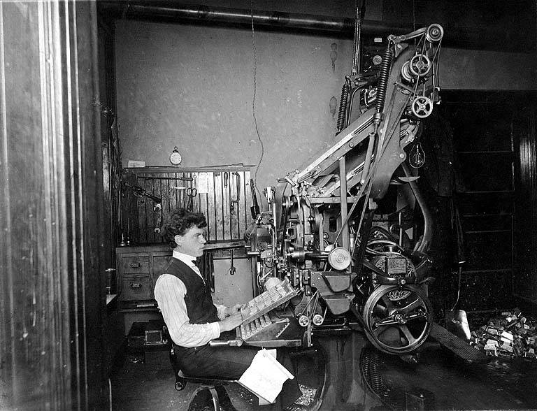A man in vintage clothing operates a large, complex mechanical typesetting machine in a dimly lit, early 20th-century print shop. Papers and mechanical parts are scattered around the workspace.