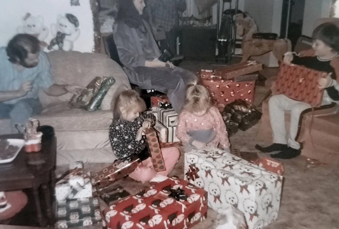 A family sits in a living room, surrounded by wrapped presents. Two children eagerly open gifts on the floor, while adults watch and unwrap presents on a couch. The atmosphere appears festive and joyful.