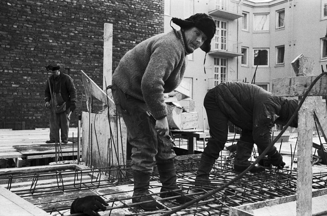 Three construction workers in warm clothing work on a building site with exposed rebar and wooden planks. One worker looks toward the camera while the others focus on their tasks. Apartments are visible in the background.
