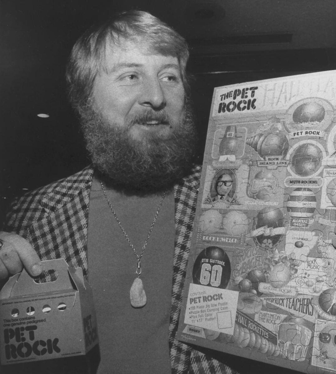 A bearded man in a plaid jacket holds a small Pet Rock box in one hand and a colorful Pet Rock manual in the other, smiling at the camera.