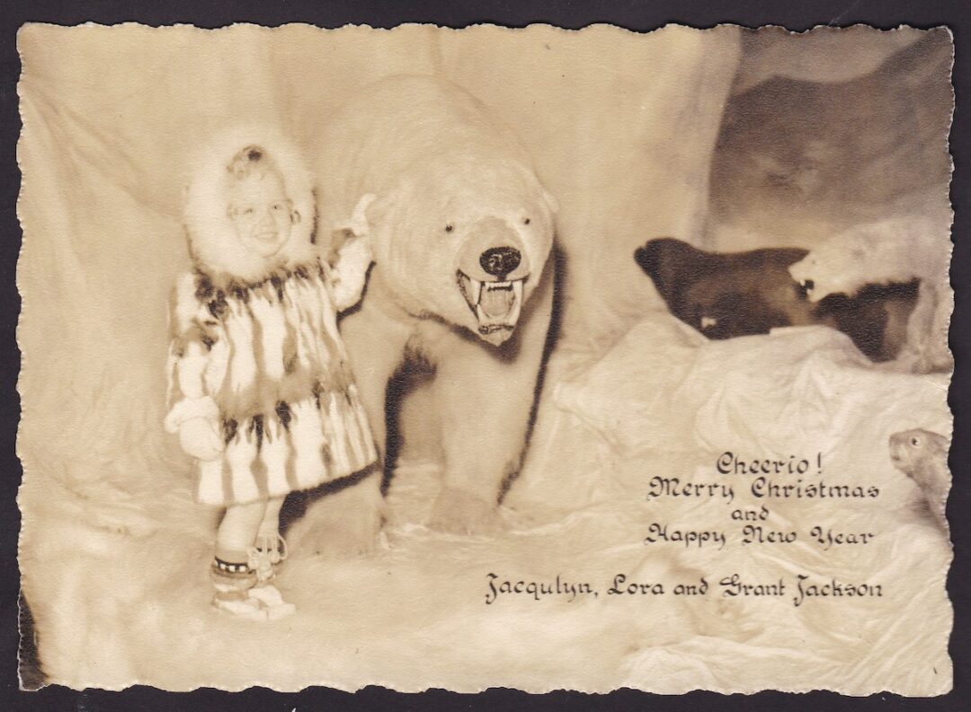 A sepia-toned photo of a smiling child in a fur-trimmed coat standing beside a taxidermy polar bear. The text reads, "Cheerio! Merry Christmas and Happy New Year, Jacqulyn, Lora and Grant Jackson.