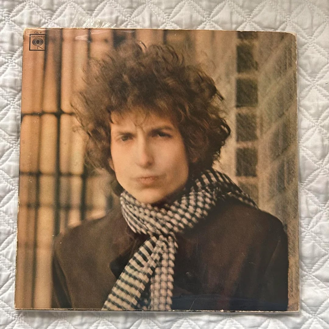 A vinyl record cover featuring a young man with curly hair wearing a checkered scarf and dark coat, standing in front of a blurred outdoor background with brick and metal bars.