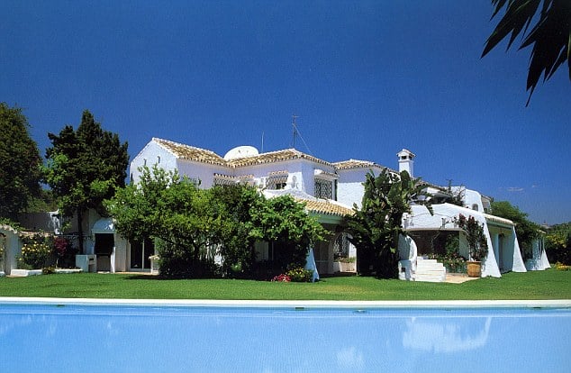 A large white villa with a tiled roof surrounded by lush green trees and grass, with a swimming pool in the foreground and a clear blue sky overhead.
