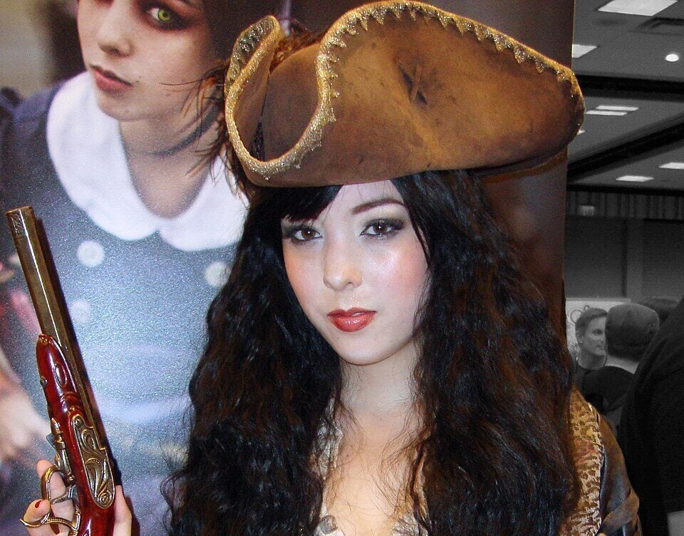 A woman in a detailed pirate costume holds a pistol and sword. She wears a large tricorn hat, a brown jacket with gold trim, a red sash, and has long dark curly hair. A blurred poster is visible in the background.