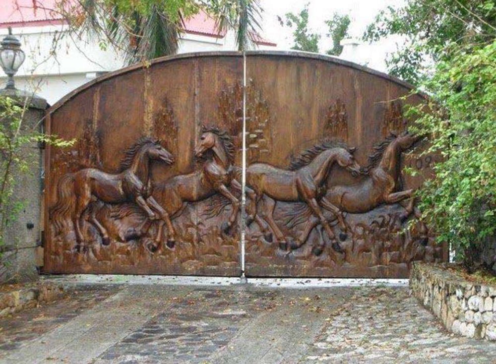 A large metal gate featuring an intricate relief of running horses, surrounded by stone walls and lush greenery, with a house and red roof partially visible behind the gate.
