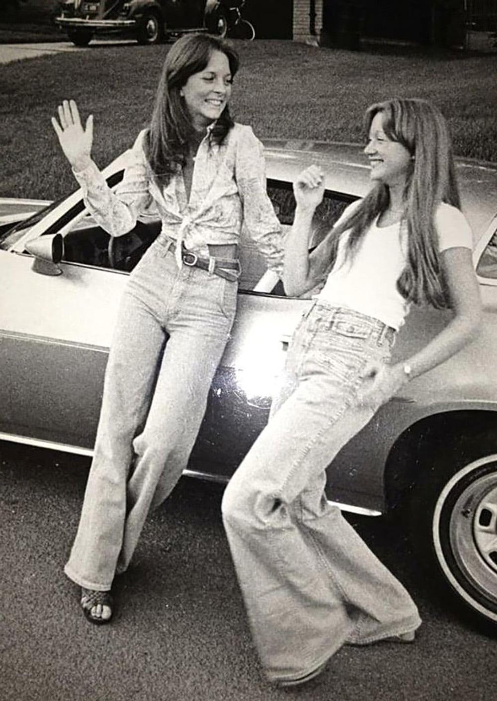 Two women in 1970s fashion, with flared jeans and long hair, smile and pose while leaning against a vintage car on a street. One woman waves, and both appear to be enjoying themselves.