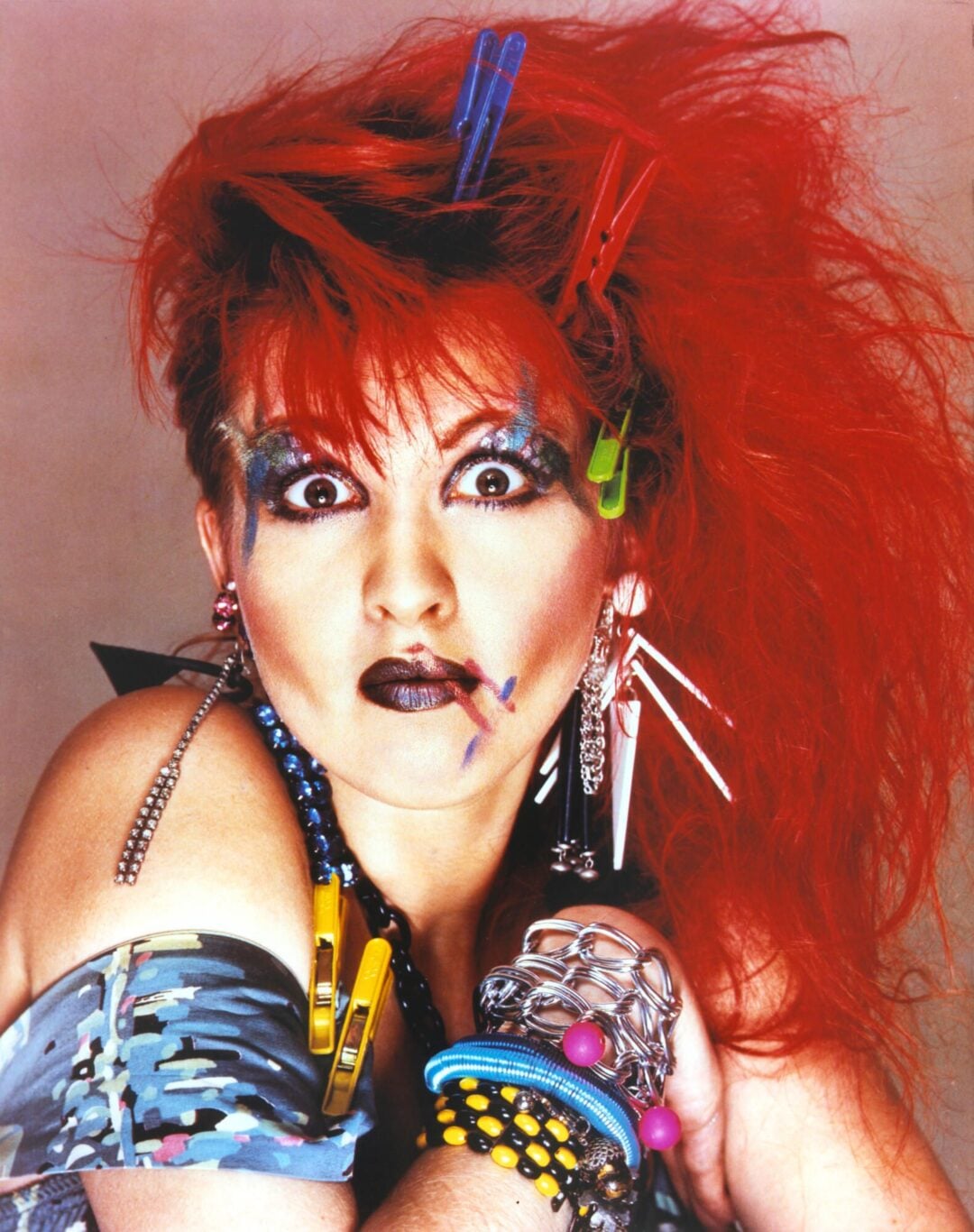 A person with bright red, teased hair styled with colorful clothespins, bold and dramatic makeup, wearing chunky bracelets, layered jewelry, and patterned off-shoulder clothing, poses with a surprised expression.