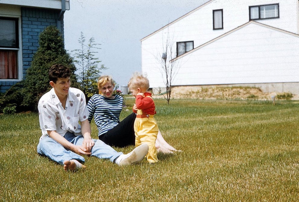 A man, woman, and toddler sit and play together on a grassy lawn in front of suburban houses on a sunny day. The adults are smiling while the toddler stands between them.