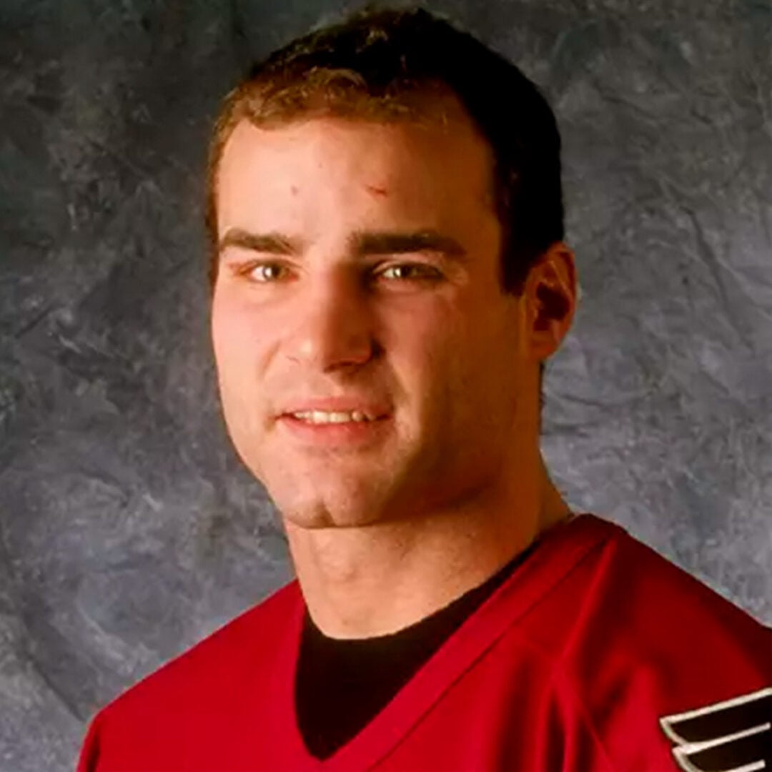 A young man with short brown hair wearing a red sports jersey stands in front of a blue-gray textured background, looking at the camera with a slight smile.