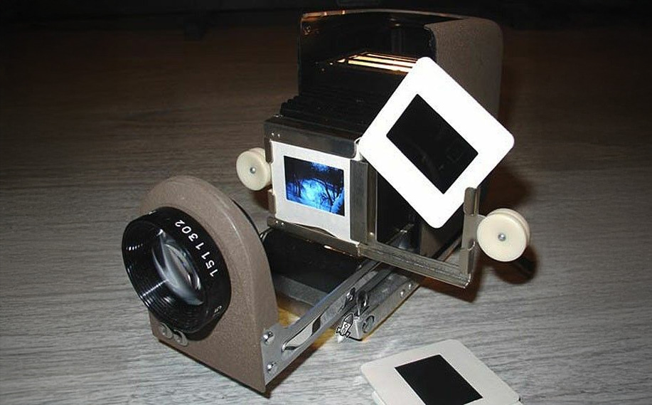 A vintage slide projector displays a photographic slide, with one slide inserted and another held nearby, on a wooden surface. The projector’s lens and internal light source are visible.