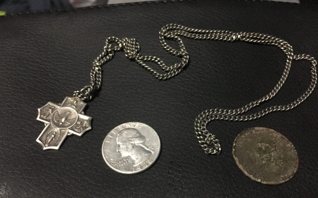 A silver cross pendant on a chain, a U.S. quarter, and a worn, round coin rest on a textured black surface. The cross pendant features engraved details and a depiction of a figure.