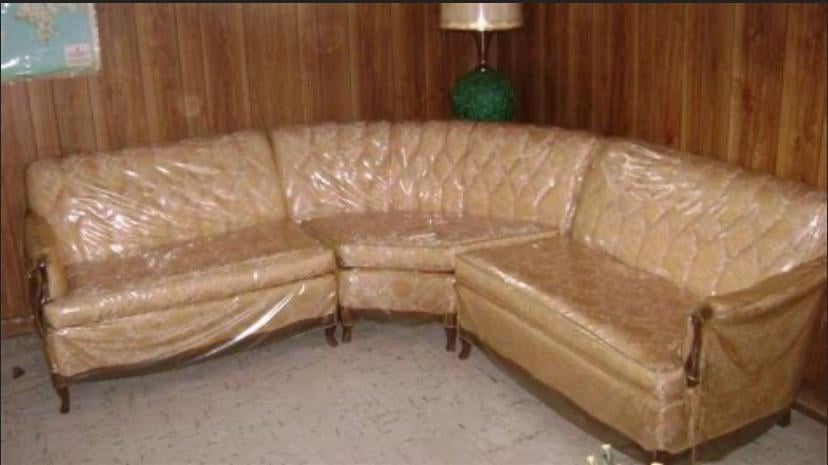 A beige, vintage sectional sofa covered in clear plastic sits in a wood-paneled room with a floor lamp and tiled floor.