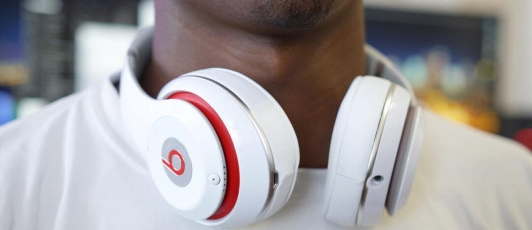 A close-up of a person wearing white and red over-ear headphones around their neck, with part of their chin and a white shirt visible. The background is slightly blurred.