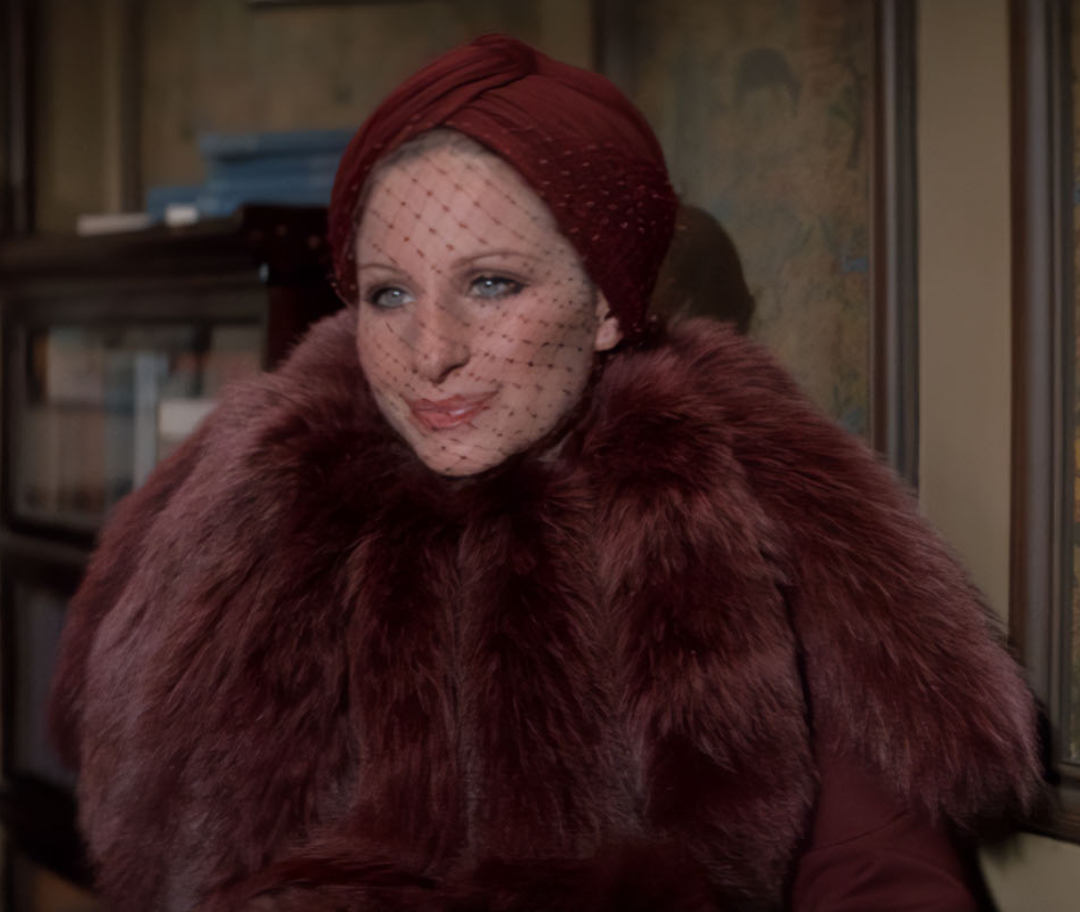 A person wearing a deep red turban, black dotted veil, and a large, plush maroon fur stole sits indoors in a vintage setting.